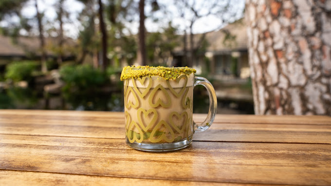 A pistachio latte with heart latte art and a dusted green rim, on a courtyard table beside the pond